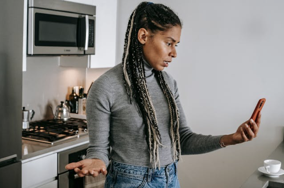 A woman with long braids, wearing a grey turtleneck and jeans, stands in a kitchen holding a phone and gesturing with her left hand, appearing frustrated or confused during a call.