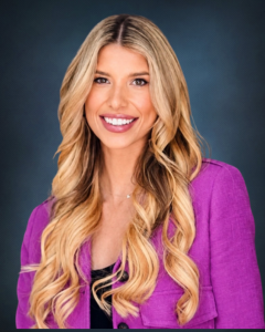 Smiling woman with long, wavy blonde hair wears a bright purple blazer over a black top, posing against a dark blue, textured background.