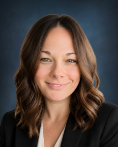 Woman with shoulder-length brown hair, wearing a black blazer and white top, smiling in front of a dark blue background.