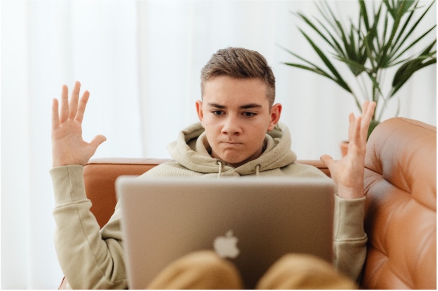 A young person in a hoodie sits on a couch, raising both hands in frustration while looking at a laptop screen, with a plant in the background.
